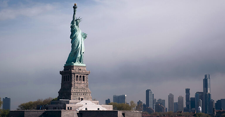 Die Freiheitsstatue auf Liberty Island vor der Skyline von New York unter bewölktem Himmel.