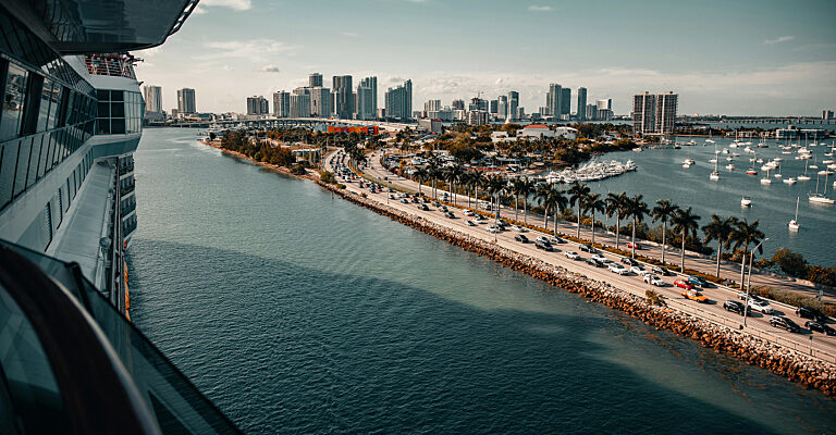 Blick von einem Kreuzfahrtschiff auf die Skyline von Miami mit palmengesäumter Uferstraße und Yachthafen.